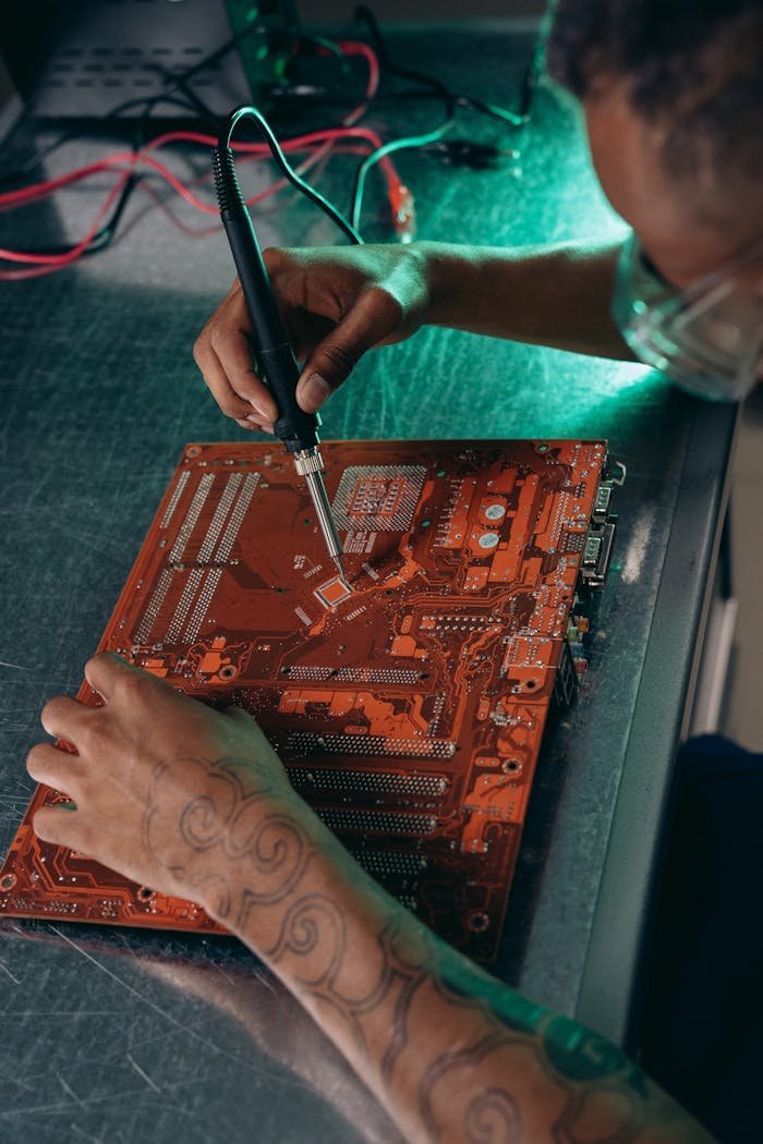 why-choose-us-02 Tattooed engineer working on a motherboard with a soldering iron in a tech workshop.